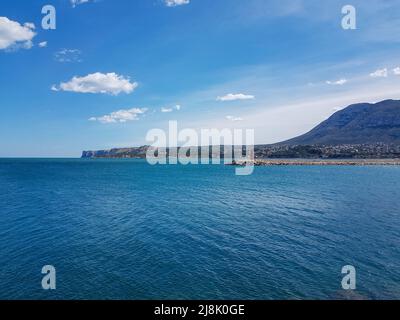 Kleiner Leuchtturm an der Hafenmauer, um den Eingang zum Hafen von Denia zu signalisieren, mit großen Küstenbergen im Hintergrund Stockfoto