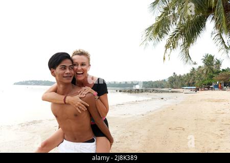 Frau lacht, während sie einen jungen Mann am Strand mit Huckepack anstößt Stockfoto