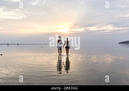 Multiethnisches Paar, das während des Sonnenuntergangs die Hände in der Mitte des Meeres hält Stockfoto