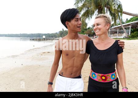 Lächelndes multikulturelles Paar, das an einem tropischen Strand entlang läuft Stockfoto