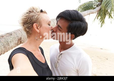 Multirassische Liebhaber küssen sich und machen ein Selfie am Strand Stockfoto