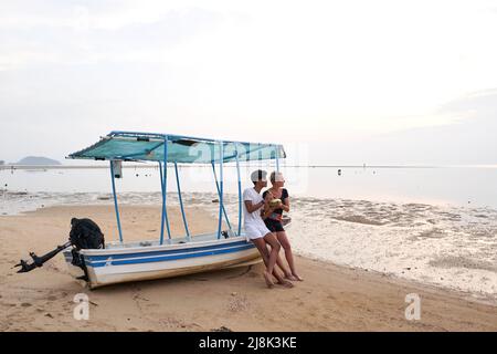 Ein Paar sitzt auf einem Boot, das im Sand eines Strandes festgemacht ist, während es eine Kokosnuss trinkt Stockfoto