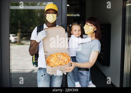 Schöne junge Frau in Gesichtsmaske hält ihre Tochter in den Armen, öffnet Türen ihres Hauses und trifft Delivery Mann, der ihren Karton mit Pizza aus dem Restaurant gibt. Stockfoto