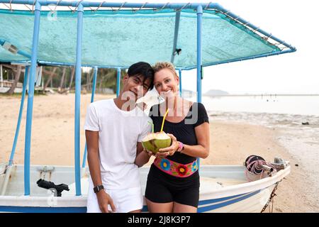 Multikulturelles Paar, das am Strand Kokosnuss trinkt und die Kamera anschaut Stockfoto