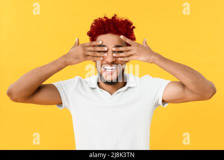 Stilvoller junger Mann mit leuchtend roten lockigen Haaren, die die Augen mit den Händen auf gelbem Hintergrund bedecken Stockfoto