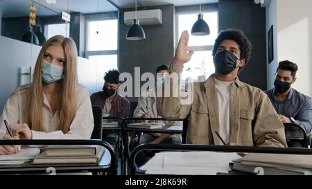 Junge afroamerikanische Studentin in medizinischer Maske sitzt am Schreibtisch unter multiethnischen Klassenkameraden wirft die Hand und stellt dem Lehrer während des Gruppenseminars Fragen Stockfoto