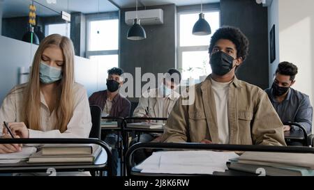 Junge afroamerikanische Studentin in medizinischer Maske sitzt am Schreibtisch unter multiethnischen Klassenkameraden wirft die Hand und stellt dem Lehrer während des Gruppenseminars Fragen Stockfoto