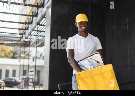 Lebensmittelverschiffung, Beruf und Menschen Konzept - glücklich lächelnder afrikanischer Lieferer mit gelber wärmegedämmter Tasche in der Nähe des modernen Gebäudes. Stockfoto