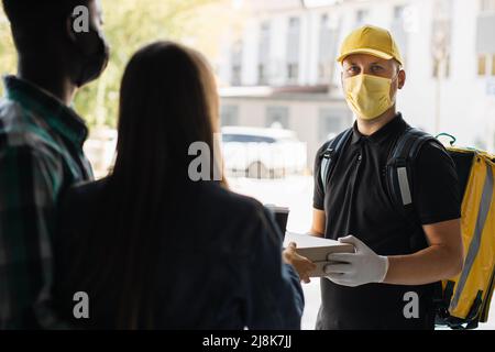 Smillender Liefermann in gelber Gesichtsmaske, einheitliche Kappe, die Pappkartons mit Pizza und Kaffee an den attraktiven internationalen Familienempfänger liefert. Kurierservice-Konzept. Stockfoto