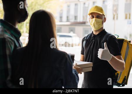 Smillender Liefermann in gelber Gesichtsmaske, einheitliche Kappe, die Pappkartons mit Pizza und Kaffee an den attraktiven internationalen Familienempfänger liefert. Kurierservice-Konzept. Stockfoto