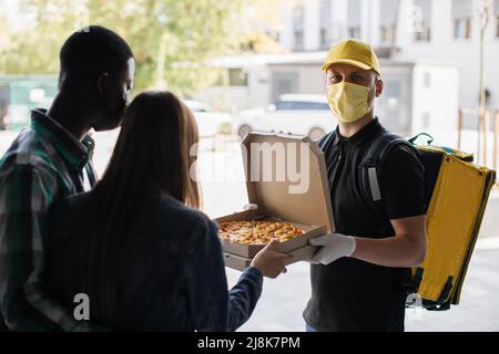 Schöne junge Frau in Gesichtsmaske steht mit ihrem Mann, öffnet Türen ihres Hauses und trifft Delivery Mann, der ihren Karton mit Pizza aus dem Restaurant gibt. Stockfoto