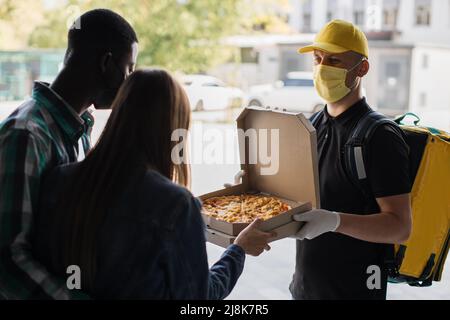 Smillender Liefermann in gelber Gesichtsmaske, einheitliche Kappe, die Pappkartons mit Pizza und Kaffee an den attraktiven internationalen Familienempfänger liefert. Kurierservice-Konzept. Stockfoto