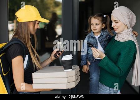 Lieferung von Lebensmitteln an eine schöne muslimische Frau mit kleiner Tochter. Ein junger Kurier liefert einen Auftrag an einen Kunden. Bestellen Sie Lebensmittel und Fast Food online. Islamische Frau zahlt per Kreditkarte, Online-Zahlung. Stockfoto