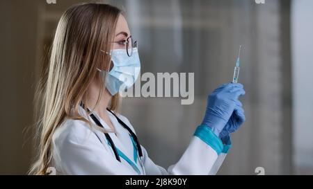Ärztin trägt eine Gesichtsmaske und einen medizinischen Kittel, der eine Spritze für die Injektion vorbereitet covid19 Impfung. Krankenschwester mit flüssiger Medizin Stockfoto