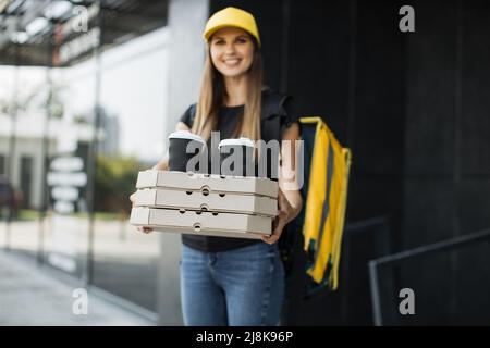 Junge glückliche kaukasische Lieferfrau mit gelbem Thermo-Box-Rucksack und Kappe. Kurier liefert Boxen mit heißer Pizza und Kaffee. Service-Konzept für schnelle Lieferung. Stockfoto
