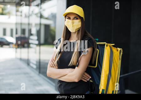 Lieferung von Lebensmitteln, Porträt einer kaukasischen Frau mit Gesichtsschutzmaske, die während einer Pandemie der Covid-19 mit gelbem Rucksack arbeitete. Schneller Service, Arbeitskonzept für Ausländer, einfacher Beruf. Stockfoto