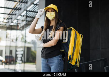 Porträt der Lieferung Frau mit gelber Kappe in Gesichtsmaske und thermischen Rucksack Blick auf die Kamera. Fröhlicher Kurier, der Fast Food zum Haus oder Büro des Kunden liefert. Express-Lieferung. Stockfoto