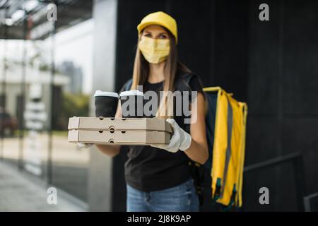 Porträt der Lieferung Frau mit gelber Kappe in Gesichtsmaske und thermischen Rucksack Blick auf die Kamera. Fröhlicher Kurier, der Fast Food zum Haus oder Büro des Kunden liefert. Express-Lieferung. Stockfoto