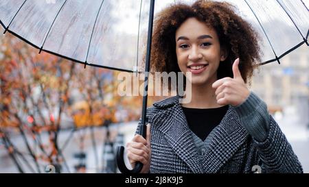 Afrikanische glücklich erfolgreiches Mädchen lockig multirassische Frau mit transparentem Regenschirm posiert in Herbststadt im Regen Blick auf Kamera zeigt Daumen nach oben Stockfoto