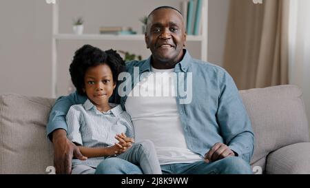 afroamerikanische Familie auf dem Sofa sitzend kleines Mädchen winkt hallo Gruß erwachsenen Vater umarmt Tochter im Gespräch an der Kamera lächelnd zeigt gesundes Weiß Stockfoto