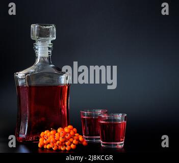 Rowan-Likörlikör, starker hausgemachter Alkohol in einer Glasdekanter und zwei Gläser auf schwarzem Hintergrund, neben einem Haufen reifer Beeren. Nahaufnahme. Stockfoto