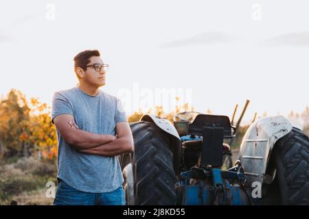 Lateinischer Bauer mit gekreuzten Armen, der neben einem alten Traktor inmitten seines Ackerlandes steht. Stockfoto