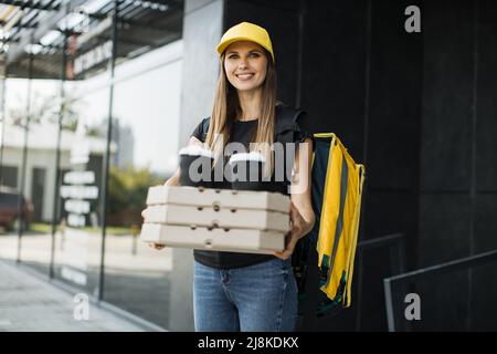 Junge glückliche kaukasische Lieferfrau mit gelbem Thermo-Box-Rucksack und Kappe. Kurier liefert Boxen mit heißer Pizza und Kaffee. Service-Konzept für schnelle Lieferung. Stockfoto