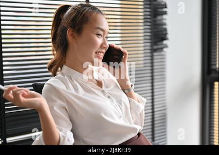 Fröhliche asiatische Geschäftsfrau oder Arbeiterin unterhalten sich gerne während der Mittagspause im Büro mit ihrer Freundin am Telefon. Stockfoto