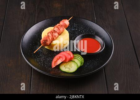 Gebratene Wurst auf Spieß mit Kartoffelpüree und Scheiben frischer Tomaten und Gurken Stockfoto