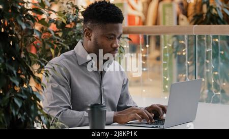 Fokussierter junger afroamerikanischer männlicher Freiberufler, der sorgfältig auf den Laptop-Bildschirm schaut, der aus der Ferne geschäftliche E-Mails schreibt und Artikel chattet Stockfoto