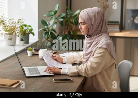 Seriöse junge Frau mit Hijab sitzt am Schreibtisch und arbeitet allein an einem neuen Geschäftsprojekt im modernen Büro Stockfoto