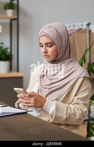 Vertikales mittleres Porträt einer modernen muslimischen Frau mit ernstem Gesichtsausdruck und SMS-Nachricht auf ihrem Smartphone Stockfoto