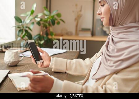 Eine moderne muslimische Geschäftsfrau mit Kopftuch sitzt am Schreibtisch und überprüft Nachrichten auf ihrem Smartphone Stockfoto
