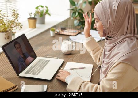 Eine fröhliche junge muslimische Frau mit Kopfbedeckung sitzt an der Rezeption und begrüßt ihren Kollegen im Internet Stockfoto