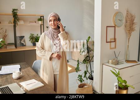 Horizontale Aufnahme einer jungen muslimischen Frau mit Kopftuch, die am Schreibtisch steht und am Telefon mit ihrer Freundin telefoniert, die wegschaut und Platz kopiert Stockfoto