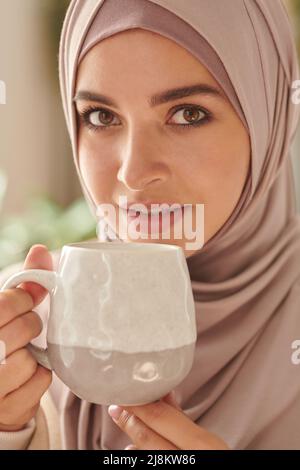 Vertikale Nahaufnahme Porträt von jungen Erwachsenen Attraktive Frau aus dem Nahen Osten trägt stilvolle pastellrosa Hijab trinken Tee Blick auf die Kamera Stockfoto