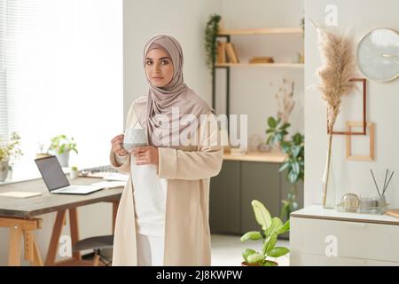 Horizontale mittlere Ling-Aufnahme einer stilvollen muslimischen Frau, die allein in einem gemütlichen Bürozimmer steht und eine Tasse Kaffee hält und Platz zum Kopieren bietet Stockfoto