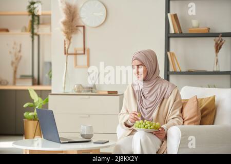Fröhliche junge Muslin-Frau mit Hijab, die in ihrem Wohnzimmer auf einem Stuhl sitzt und Film auf einem Laptop anschaut und frische Trauben isst Stockfoto