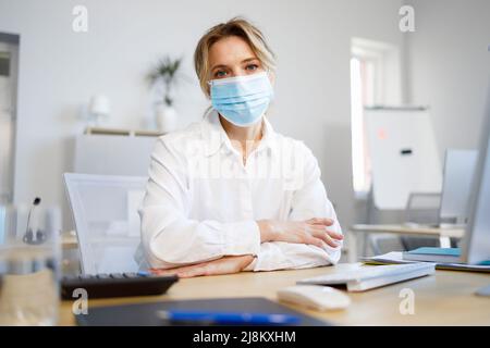 Hübsche Geschäftsfrau, die während einer Pandemie im Büro sitzt und eine Maske trägt Stockfoto