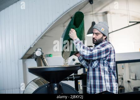 Männlicher Kaffeeröster, der rohe Kaffeebohnen in eine moderne Röstermaschine gießt Stockfoto