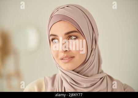 Nahaufnahme des Kopfes und der Schultern einer jungen erwachsenen muslimischen Frau, die einen blaßrosa Hijab trägt und wegschaut Stockfoto