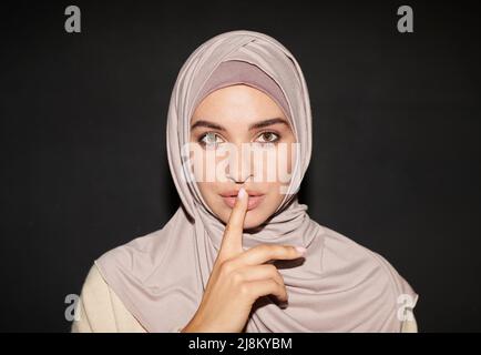 Waagerechte Kopf- und Schulteraufnahme einer modernen muslimischen Frau, die Hijab trägt, mit Vorderfinger über den Lippen, um eine Geste des Schweigens zu machen Stockfoto