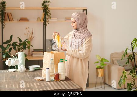 Horizontale, mittellange Aufnahme einer jungen muslimischen Frau mit Kopftuch, die am Tisch stand und das ihr gelieferte Essen herausnahm Stockfoto