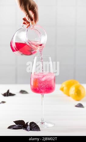 Eine Frauenhand gießt erfrischende rote Basilikumlimonade aus einem Krug in ein Glas. Sommergetränke. Stockfoto