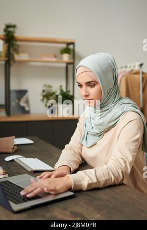 Vertikales Porträt einer schönen jungen Frau mit hellblauem Hijab, die zu Hause am Tisch sitzt und am Laptop arbeitet Stockfoto