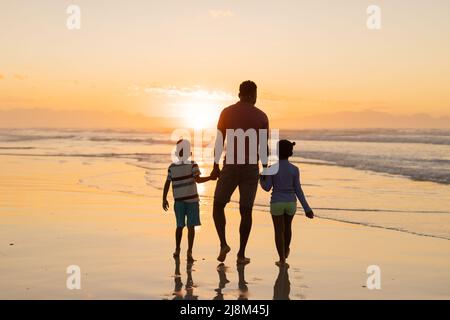 Rückansicht des afroamerikanischen jungen Vaters, der beim Strandspaziergängen die Hände von Sohn und Tochter hält Stockfoto