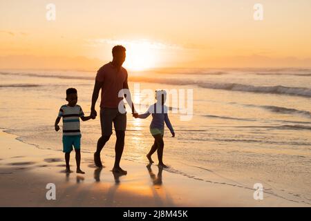 afroamerikanischer junger Vater hält die Hände von Sohn und Tochter, während er am Strand gegen den Himmel läuft Stockfoto