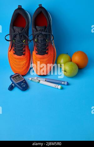 Orangefarbene Turnschuhe, grüne Äpfel, Orangen, Glucometer und Insulin-Spritzenstifte auf blauem Hintergrund mit Kopierfläche. Therapie gegen Insulinresistenz und d Stockfoto