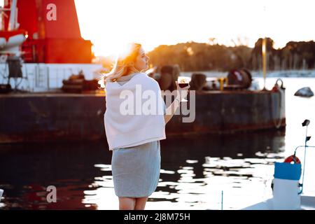 Blonde Frau schaut weg mit einem Glas roter Rebe wartet auf Pier. Urlaub auf Reisen. Romantischer Blick auf den Sonnenuntergang Stockfoto
