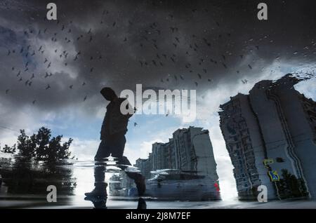 Verschwommene Silhouette der Spiegelung einer Person, die an regnerischen Tagen allein auf dem nassen Bürgersteig der Stadt läuft. Vögel fliegen über den Himmel. Abstrakte Fotografie. Stockfoto